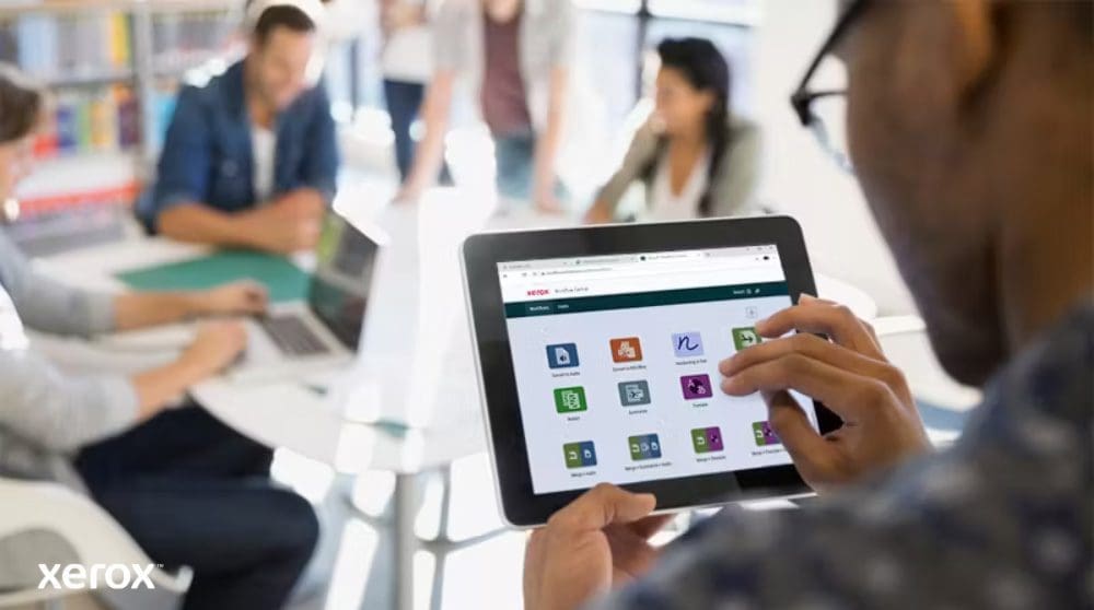 Une personne tient une tablette affichant l'interface Xerox Workflow Central avec diverses ic&ocirc;nes d'application, tandis que d'autres personnes travaillent dans l'arri&egrave;re-plan flou, mettant en &eacute;vidence l'acc&egrave;s mobile aux solutions de flux de travail.